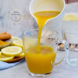 Vertiendo té de cúrcuma caliente en una taza de cristal, con limones en rodajas, ramas de canela y cúrcuma en polvo en el fondo, añadiendo un toque visual de frescura.