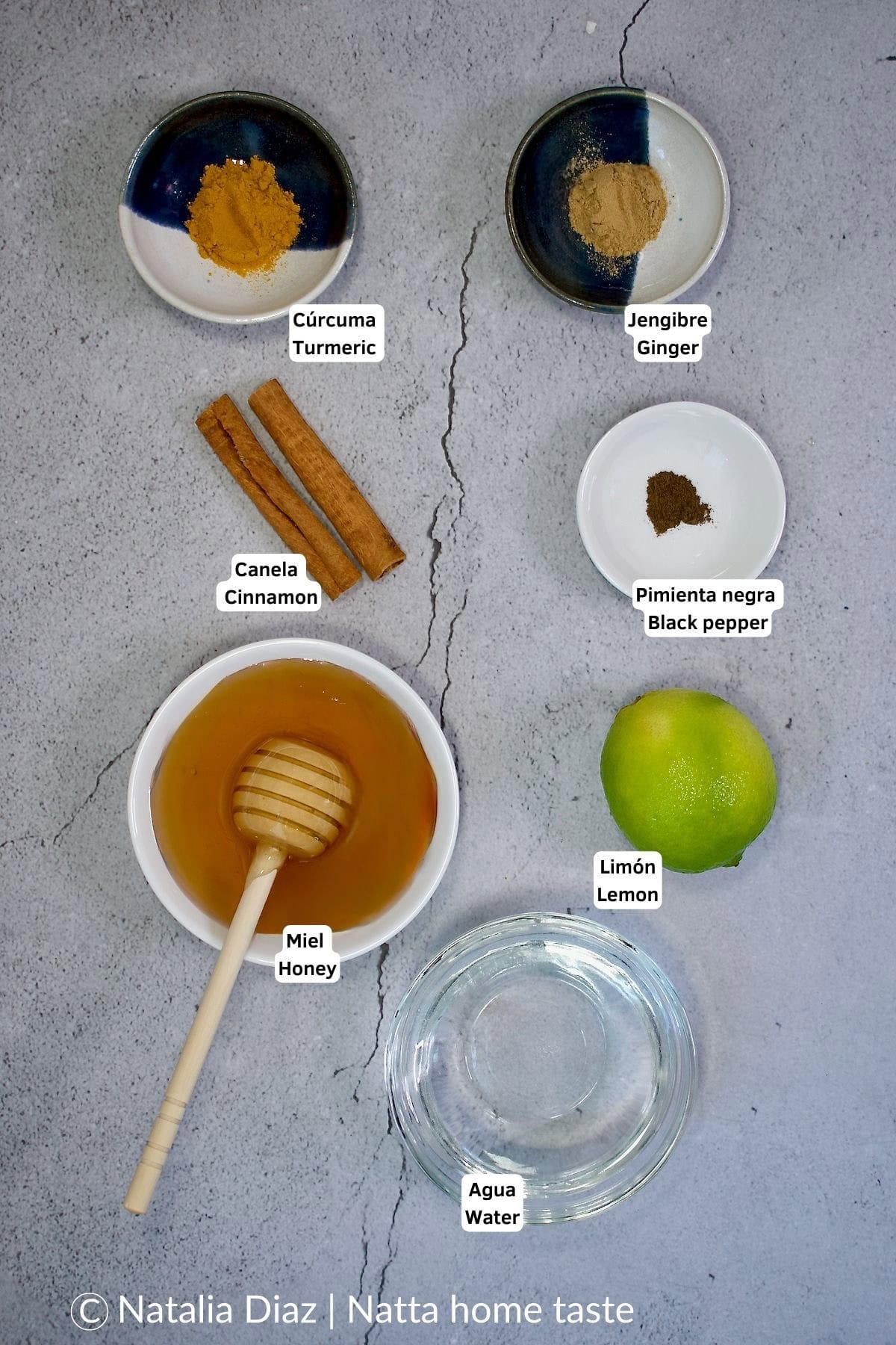 Ingredientes necesarios para la receta de té de cúrcuma, incluyendo cúrcuma en polvo, jengibre, canela, limón, pimienta negra, miel y agua, dispuestos en platos pequeños sobre una superficie gris.