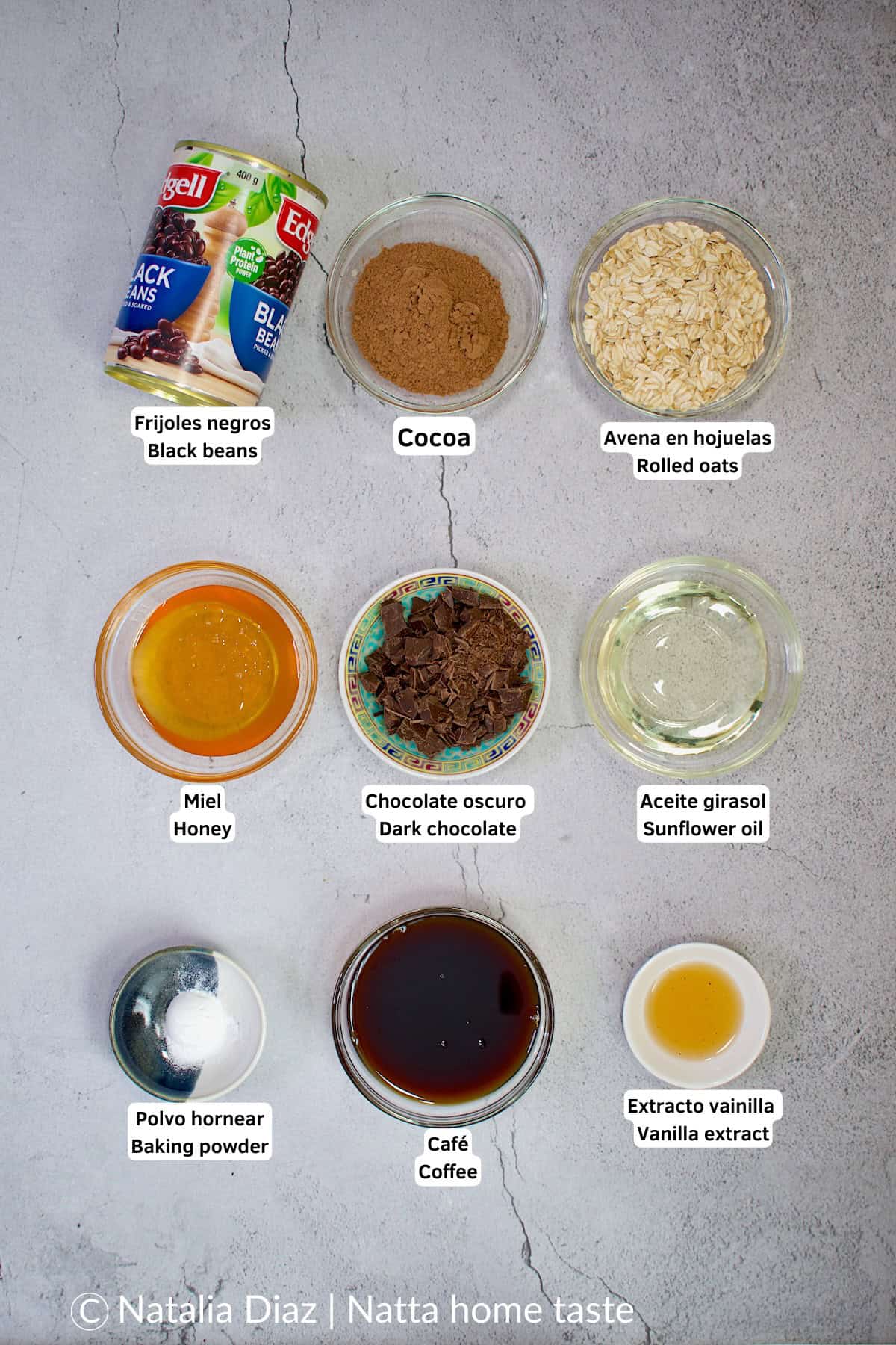 Ingredientes para hacer brownies sin huevo: frijoles negros enlatados, cocoa en polvo, avena en hojuelas, miel, chocolate oscuro troceado, aceite de girasol, polvo para hornear, café y extracto de vainilla. Todos dispuestos en pequeños recipientes sobre una superficie gris.
