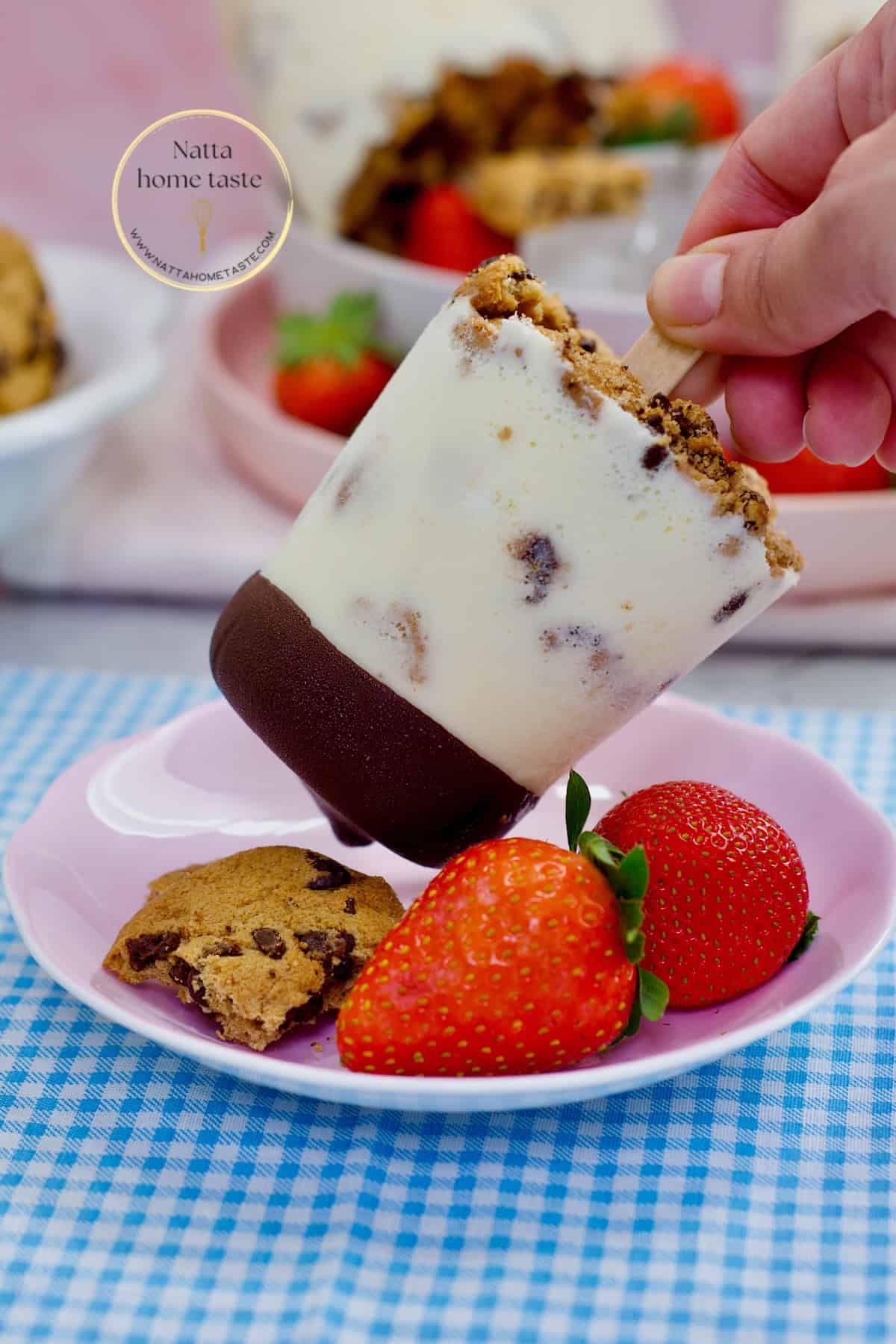 Paleta de leche cremosa con trozos de galleta y cobertura de chocolate, servida con fresas frescas y una galleta en un plato rosado.