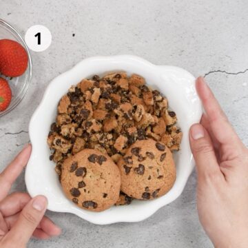 Tazón blanco con galletas con chips de chocolate partidas en trozos, listo para preparar paletas caseras. Paso 1 de la receta.