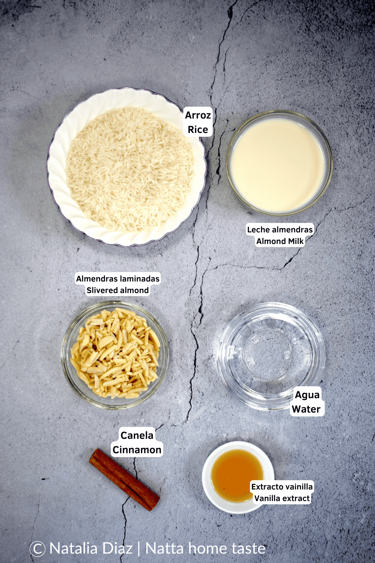 Ingredientes para hacer horchata de arroz casera: arroz crudo, leche de almendras, almendras laminadas, agua, rama de canela y extracto de vainilla, dispuestos sobre una superficie gris con etiquetas en español e inglés.