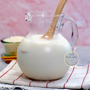 Jarra de horchata de arroz casera con hielo y cuchara de madera, sobre un paño de cocina, con arroz y canela al fondo. Bebida refrescante tipo agua fresca mexicana.