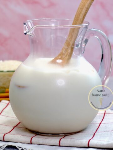 Jarra de horchata de arroz casera con hielo y cuchara de madera, sobre un paño de cocina, con arroz y canela al fondo. Bebida refrescante tipo agua fresca mexicana.
