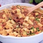 Pasta corta con carne molida en salsa de tomate, en sartén blanco; cucharón de madera levantando porción y albahaca fresca picada.