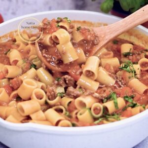 Pasta corta con carne molida en salsa de tomate, en sartén blanco; cucharón de madera levantando porción y albahaca fresca picada.