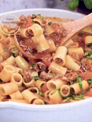 Pasta corta con carne molida en salsa de tomate, en sartén blanco; cucharón de madera levantando porción y albahaca fresca picada.
