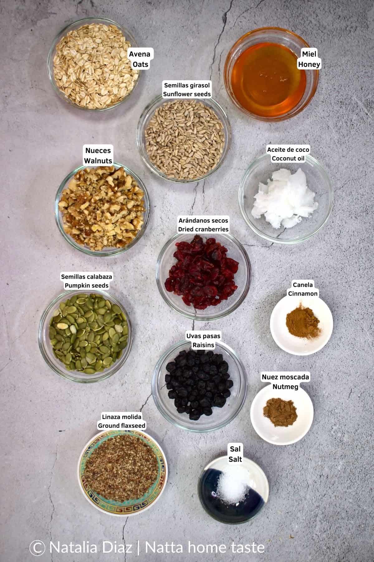 Foto cenital de los ingredientes para granola casera: avena en hojuelas, miel, aceite de coco, nueces, semillas de girasol y de calabaza, arándanos secos, uvas pasas, linaza molida, canela, nuez moscada y sal, en pequeños tazones de vidrio sobre fondo gris.