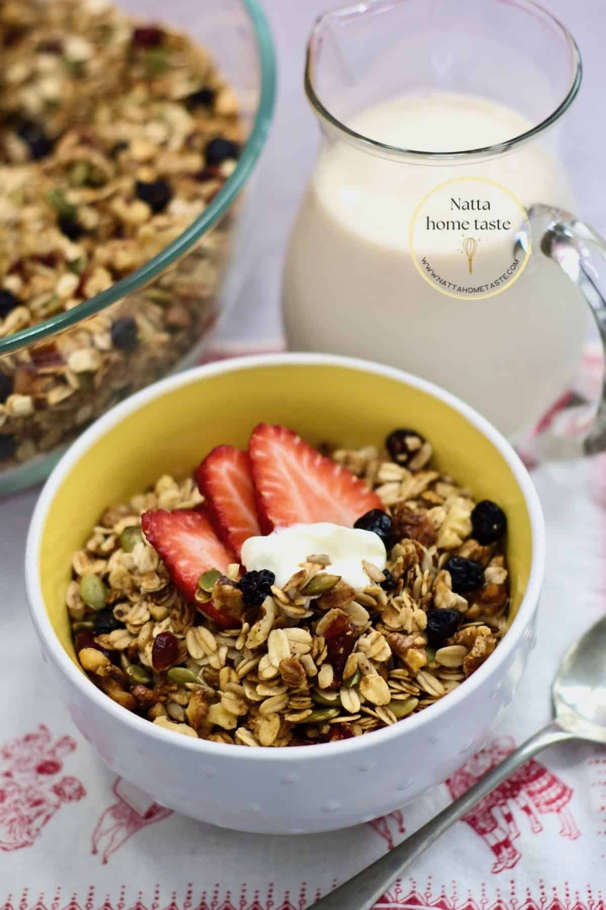 Tazón blanco con granola casera crujiente hecha en freidora de aire, servida con fresas, una cucharada de yogur, pepitas y arándanos secos; jarra de leche y cuchara al fondo.