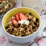 Tazón blanco con granola casera crujiente hecha en freidora de aire, servida con fresas, una cucharada de yogur, pepitas y arándanos secos; jarra de leche y cuchara al fondo.