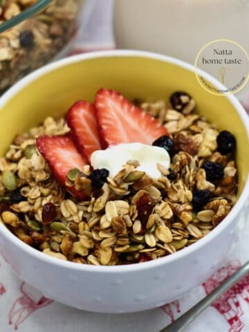 Tazón blanco con granola casera crujiente hecha en freidora de aire, servida con fresas, una cucharada de yogur, pepitas y arándanos secos; jarra de leche y cuchara al fondo.