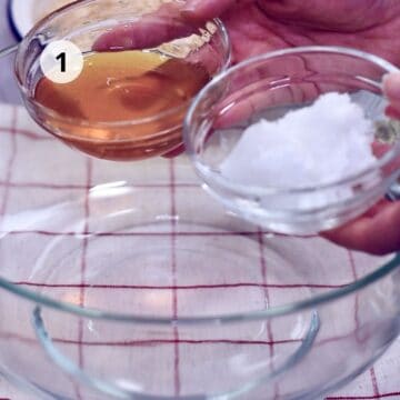 Manos sosteniendo dos tazones de vidrio, uno con miel dorada y otro con aceite de coco sólido, sobre un bol grande transparente; fondo de mantel blanco con cuadriculado rojo y círculo con número 1 indicando el paso.