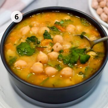 Paso 6: tazón negro con sopa de garbanzos lista para servir, con cilantro fresco y trozos de zanahoria; una cuchara dentro.