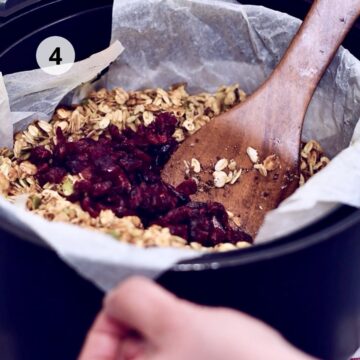 Paso 4: en la canasta de una freidora de aire forrada con papel para hornear, una cuchara de madera mezcla la granola mientras se incorporan arándanos secos; mano sujetando el borde y círculo con número 4.