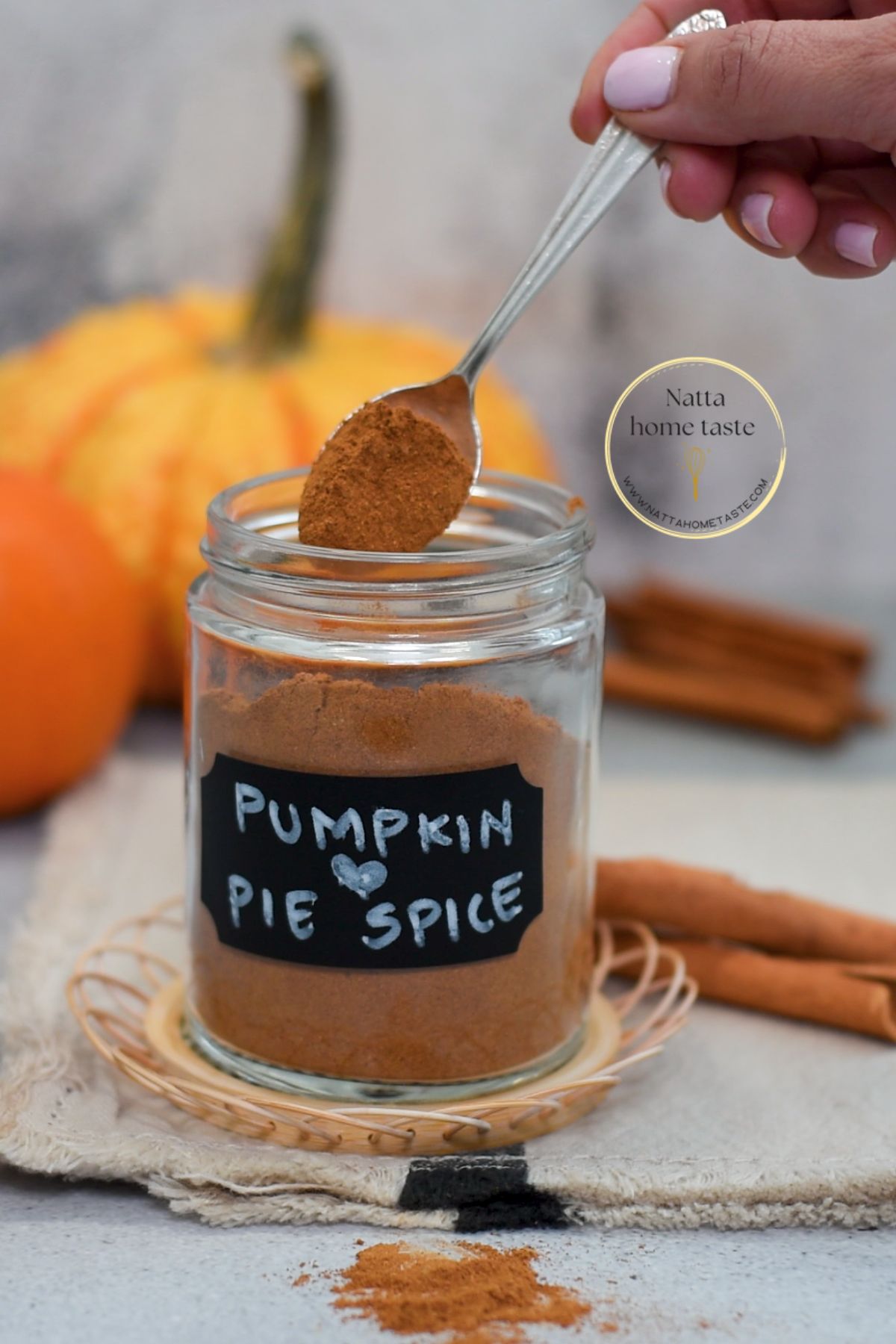 Frasco de vidrio con mezcla casera de especias para tarta de calabaza, con etiqueta “Pumpkin Pie Spice” y fondo de calabazas y ramas de canela sobre una tela beige.
