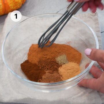 Mezcla de especias para pastel de calabaza en proceso, con canela, jengibre, clavo y nuez moscada en un bowl de vidrio, mientras se baten con un batidor de mano.