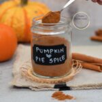 Frasco de vidrio con mezcla casera de especias para tarta de calabaza, con etiqueta “Pumpkin Pie Spice” y fondo de calabazas y ramas de canela sobre una tela beige.