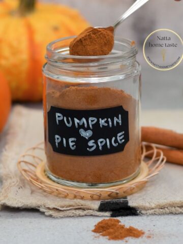 Frasco de vidrio con mezcla casera de especias para tarta de calabaza, con etiqueta “Pumpkin Pie Spice” y fondo de calabazas y ramas de canela sobre una tela beige.