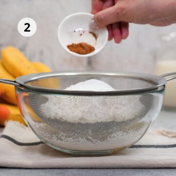 Mano añadiendo canela desde un pocillo a un colador sobre un bol de vidrio mientras se tamizan los ingredientes secos; etiqueta “2” en la esquina y frutas desenfocadas al fondo sobre un paño de cocina.