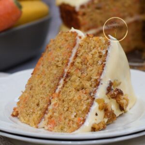 Pastel de zanahoria y plátano de 2 capas con miga húmeda, frosting de queso crema con efecto “drip” y nueces; rebanada en primer plano sobre plato blanco.
