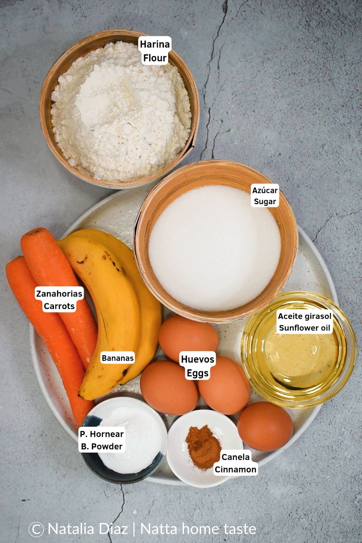 Ingredientes para pastel de zanahoria y plátano: harina, azúcar, aceite de girasol, huevos, bananas, zanahorias, polvo de hornear y canela, dispuestos en plato con rótulos en español e inglés. Vista cenital.