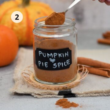 Frasco de vidrio con mezcla casera de especias para pastel de calabaza, rotulado “Pumpkin Pie Spice”, mientras se coloca una cucharadita de la mezcla dentro del frasco, con calabazas y ramas de canela al fondo.