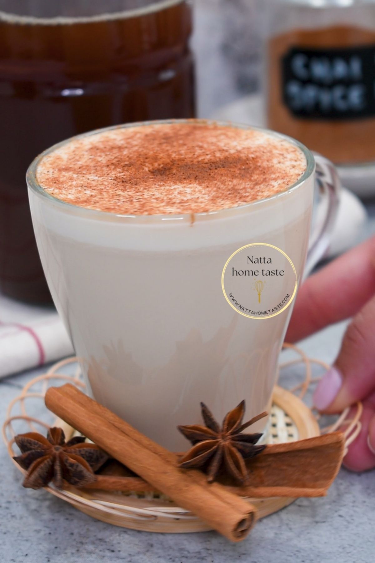 Taza de té chai casero con leche espolvoreada con canela, decorada con ramas de canela y anís estrellado al frente. Al fondo se ve el concentrado de té chai en frasco y un frasco pequeño con mezcla de especias.