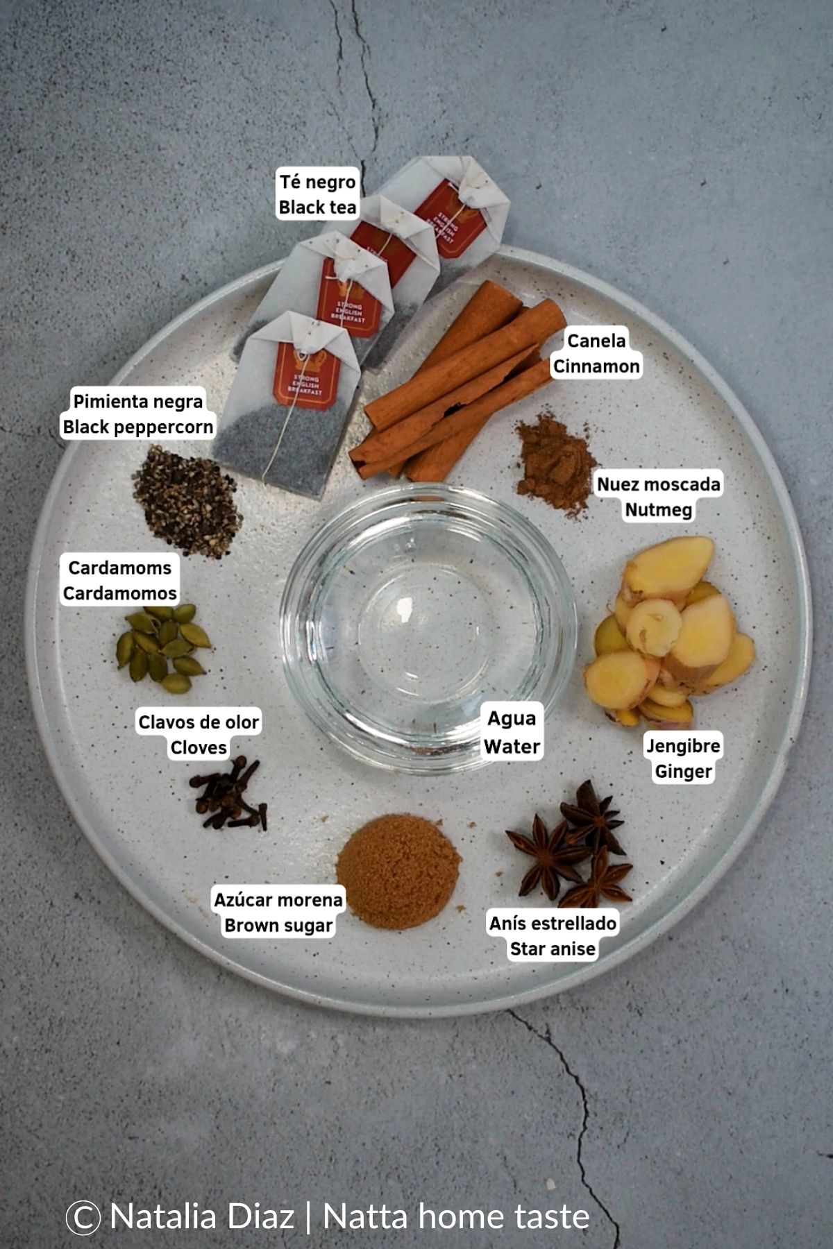 Plato blanco con los ingredientes del té chai dispuestos en secciones: bolsitas de té negro, canela en rama, pimienta negra molida, cardamomos, clavos de olor, azúcar morena, anís estrellado, jengibre fresco en rodajas, nuez moscada y un pequeño recipiente con agua.