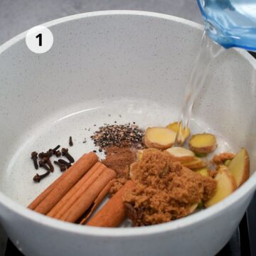Olla blanca con especias enteras: canela, clavos, pimienta negra, jengibre fresco y azúcar morena, mientras se vierte agua caliente para preparar la infusión.
