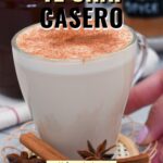 Taza de té chai casero con leche espolvoreado con canela, acompañada de astillas de canela y anís estrellado sobre una base tejida, con frascos de especias al fondo.
