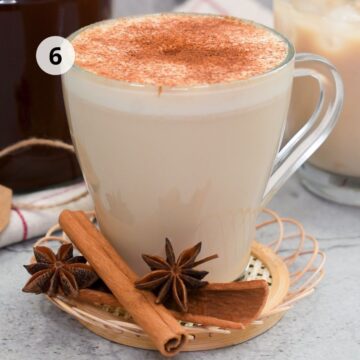 Taza transparente con bebida caliente con leche espolvoreada con canela, servida sobre una base tejida con ramas de canela y anís estrellado al frente; al fondo se observa un vaso con una bebida fría y una jarra de concentrado.