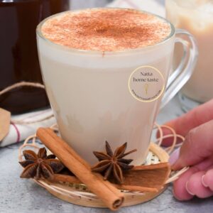 Taza de té chai con leche espolvoreada con canela, servida sobre una base tejida con ramas de canela y anís estrellado al frente, junto a una mano sosteniendo el platito.