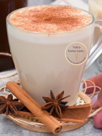 Taza de té chai con leche espolvoreada con canela, servida sobre una base tejida con ramas de canela y anís estrellado al frente, junto a una mano sosteniendo el platito.