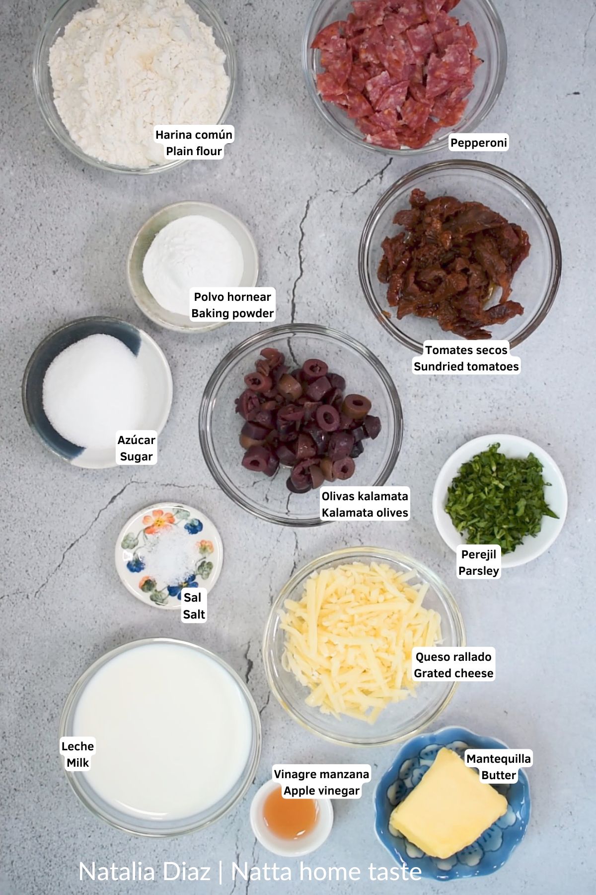 Ingredientes organizados en recipientes sobre una superficie clara: harina de trigo, queso rallado, aceitunas, pepperoni, tomates secos, perejil, leche, mantequilla y otros ingredientes para preparar un pan casero.
