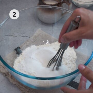 Tazón de vidrio con harina, azúcar y polvo de hornear mientras se mezclan con un batidor de mano.